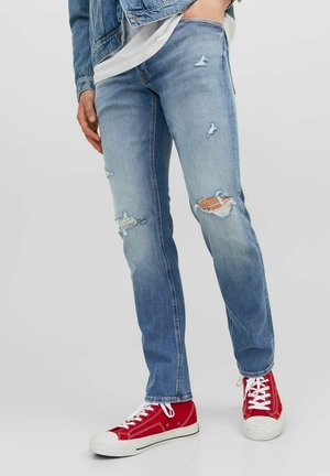 Jeans de mezclilla azul claro con parches desgastados y dobladillos deshilachados, combinados con zapatillas de lona rojas con cordones blancos y suelas de goma.