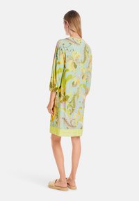 Kurzes Blumenkleid in Hellgrün mit Puffärmeln und gelbem Streifen am Saum. Hat ein farbenfrohes botanisches Muster und leichte Stoffqualität.