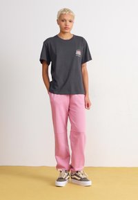 Svart bomullst-skjorte med logo, rosa joggebukser, og grå sneakers med hvite detaljer, stående på en gul overflate mot en ensfarget bakgrunn.