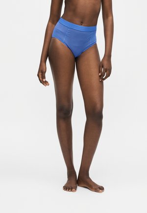 Personne debout portant un sous-vêtement bleu côtelé taille haute sur un fond uni clair, montrant le torse et les jambes nues.