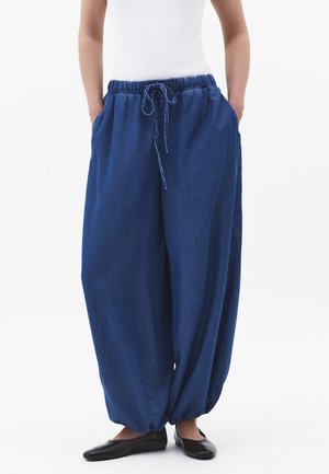 Løse mørkeblå bukser med elastik i taljen og snøre, båret med sorte flade sko og hvid ærmeløs top, hænder i lommerne.