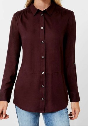 Frau trägt ein langärmliges, dunkel bordeauxrotes Hemd mit Knöpfen und Kragen sowie hellblaue Jeans vor einfachem Hintergrund.