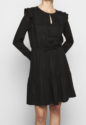 Femme portant une robe noire à manches longues avec des épaules à volants, un décolleté en forme de goutte d'eau, une taille cintrée avec un lien, et une jupe à étages.