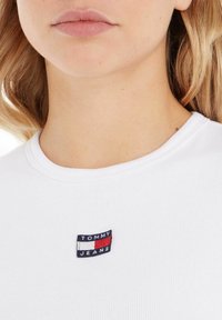 Bílý žebrovaný tričko s našitým námořnickým modrým, červeným a bílým logem "TOMMY JEANS" na levé straně hrudi.