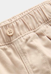 Lys beige bukser med en samlet talje, en knaplukning og synlige syninger. Stoffets tekstur er glat og blød.