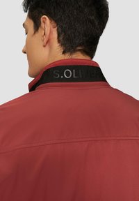 Červená bunda s čiernym golierom a logom "S.OLIVER". Hladká textúra, jednoduchý dizajn a viditeľné švy na zadnej strane.