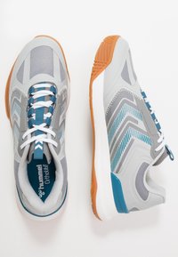 Grijze sport schoenen met blauwe accenten, textuur mesh bovenwerk, witte veters en een oranje rubberen zool. Bevat zigzag ontwerp elementen.