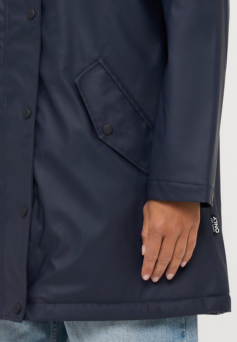 Manteau imperméable navy avec boutons-pression, une poche latérale et une texture lisse et brillante, avec une étiquette visible sur l'ourlet.
