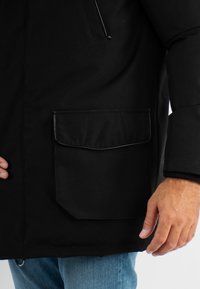 Veste noire dotée d'une grande poche avant avec un rabat, fabriquée en tissu lisse. Le style comprend une poche poitrine zippée et des poignets côtelés.