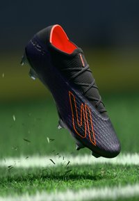 Scarpino da calcio nero con accenti arancioni, tomaia texturizzata e lacci neri, caratterizzato da un design curvo e tacchetti, catturato in aria sopra l'erba.
