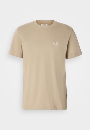 Camiseta de algodón beige con cuello redondo, mangas cortas y un pequeño parche rectangular de logo en el lado izquierdo del pecho. Textura suave.