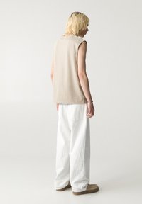 Chemise beige sans manches, coupe décontractée, associée à un pantalon blanc ample. Design simple, couture minimale, tissu texturé. Chaussures de couleur neutre.