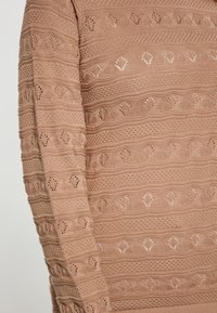 Pulover tricotat în bej moale, cu modele texturate, având forme de diamant și secțiuni subtile de țesătură deschisă pe mâneci.