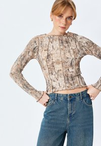 Top lungo a maniche corte con motivo a stampa serpente in tonalità beige e marrone, con orlo ondulato e tessuto testurizzato. Abbinato a jeans blu.
