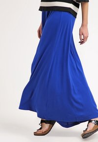 Jupe maxi bleue en tissu doux et fluide, présentant une texture lisse. Associée à des sandales noires avec une large bride et une semelle texturée.