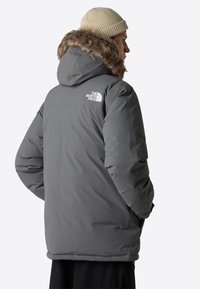 Grå isolerad jacka med fuskpälsfoder i huvan, med en logotyp på övre ryggen och en något lös passform för lätt rörelse.