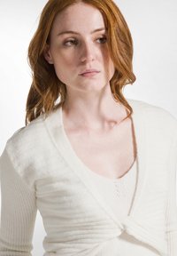 Weißer gestrickter Cardigan mit geripptem Muster und einem verdrehten Vorderteil, getragen über einem passenden Top mit rundem Ausschnitt. Weicher Stoff mit lockerem Schnitt.