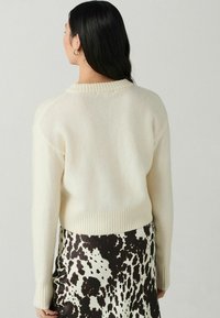 Romig gekleurd gebreid trui met een geribbelde zoom en manchetten, gedragen over een koeienprint rok. Zachte textuur, relaxed fit en ronde halslijn.