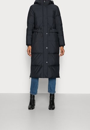 Long manteau d'hiver noir matelassé avec capuche, boutons-pression sur le devant, liens de taille réglables, porté avec un jean bleu et des bottines noires.