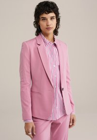Růžový blazer s špičatými klopami, na jedno knoflíček a boční kapsy, doplněný pruhovanou košilí s růžovými a bílými horizontálními pruhy.