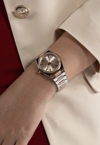 Silver and gold klocka med rund urtavla, texturerad bronstid och kristallaccenter. Metallarmband med växlande segment i guld och silver.