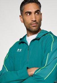 Τζελούν με κουκούλα σε τυρκουάζ χρώμα με αντανακλαστικές κίτρινες λεπτομέρειες· φερμουάρ μπροστά· ελαφρύ, υφασμάτινο με υφή· λογότυπο στο πάνω αριστερό μέρος του στήθους.