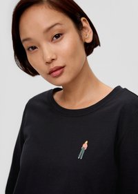 Kobieta z krótkimi ciemnymi włosami, ubrana w czarną koszulę z małą wyszywaną postacią w różowym i zielonym na piersi, na jednolitym jasnym tle.