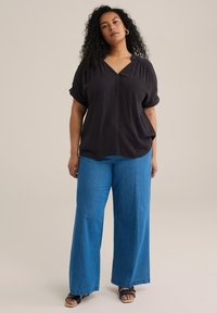 Čierny krátky rukáv top s výstrihom do V a zhrnutými ramenami, doplnený širokými modrými džínsami. Sandále s remienkami okolo členkov.