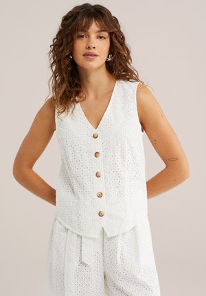 Vrouw draagt een mouwloze witte eyelet top met knopen en bijpassende shorts, staand met handen achter de rug tegen een effen achtergrond.