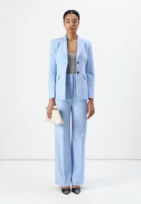 Ljust blå skräddarsydd kostym med enblads blazer och högmidjade byxor. Tillbehören inkluderar en liten krämfärgad handväska och svarta klackar.