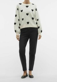 Suéter blanco con patrones de corazones negros, de mangas largas y cuello redondo, combinado con jeans negros ajustados y bailarinas negras.