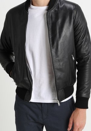 Man draagt een zwarte leren bomberjas met rits over een wit t-shirt en donkere, met patroon bedrukte broek, handen ontspannen langs het lichaam.