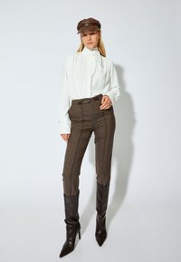 Blusa bianca con un fiocco al collo, pantaloni marroni a vita alta e stivali marroni punta a ginocchio. Un cappello marrone completa l'outfit.