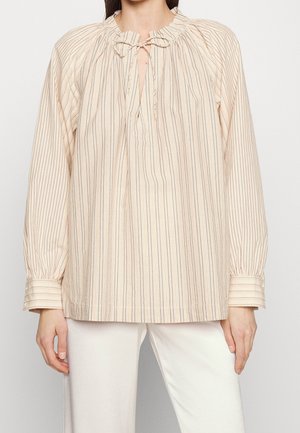 Vrouw die een beige blouse met lange mouwen draagt, voorzien van dunne verticale strepen, een geplooide halslijn en een strik aan de voorkant, gecombineerd met een witte broek.