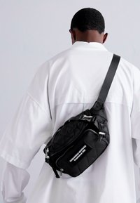 Quilted black sling bag made of nylon, featuring a silver zipper, adjustable strap, and a rectangular tag with white text.