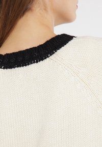 Kremowy sweter wykonany na drutach z grubym, czarnym ściągaczem. Tekstura materiału jest miękka, a w pobliżu dekoltu widać luźne szwy.