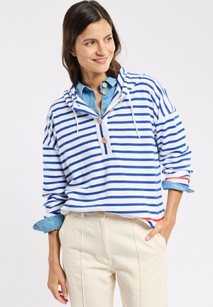 Femme souriante portant un pull rayé bleu et blanc avec des boutons en bois, une chemise en jean bleue en dessous et un pantalon beige.