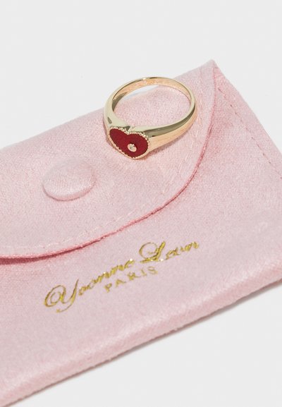 Inel de aur cu un design în formă de inimă roșie și o mică pietricică prețioasă, așezat pe un pliculeț de catifea roz cu detalii aurii.