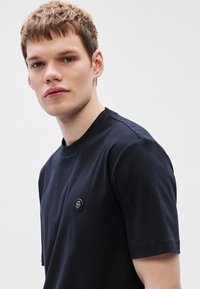 Μπλουζάκι σε σκούρο μπλε ναυτικό χρώμα με κοντά μανίκια, κατασκευασμένο από βαμβάκι, με στρογγυλή λαιμόκοψη και μικρό μαύρο λογότυπο στο αριστερό στήθος.