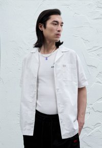 Camicia bianca a maniche corte con taschino sul petto, indossata sopra una canottiera leggera. Presenta un dettaglio con logo ed è abbinata a pantaloncini sportivi neri.