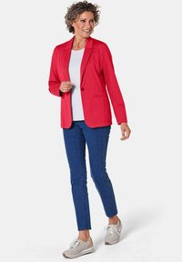 Rode blazer met revers, gecombineerd met een witte top en blauwe jeans. Accessoires omvatten lichte sneakers. Glad materiaal, slim fit.
