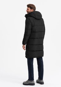 Sort puffervinterjakke med hætte, quiltet tekstur og ribbede manchetter. Bæret med mørke jeans og grå fodtøj, vist fra bagsiden.