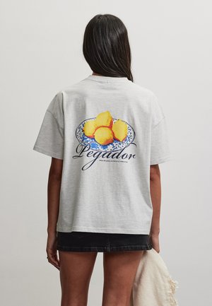 Vrouw draagt een lichtgrijs t-shirt met een blauw en geel citroenbordgrafiek en de tekst "Pegador" op de achterkant, gecombineerd met een zwarte spijkerrok.