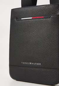 Czarna torba skórzana z teksturowanej skóry o wyraźnym kształcie, z przednią kieszenią z czerwonymi i białymi akcentami oraz srebrnym logo. Regulowany pasek.