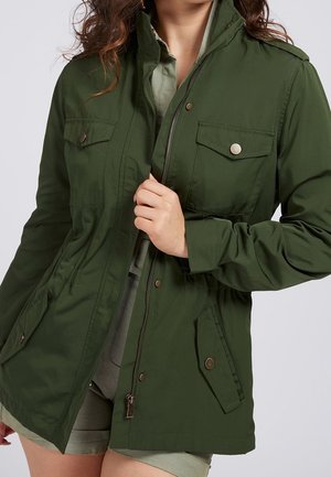 Femme portant une veste utilitaire vert olive avec poches avant et boutons-pression en laiton, partiellement zippée sur des vêtements vert clair.