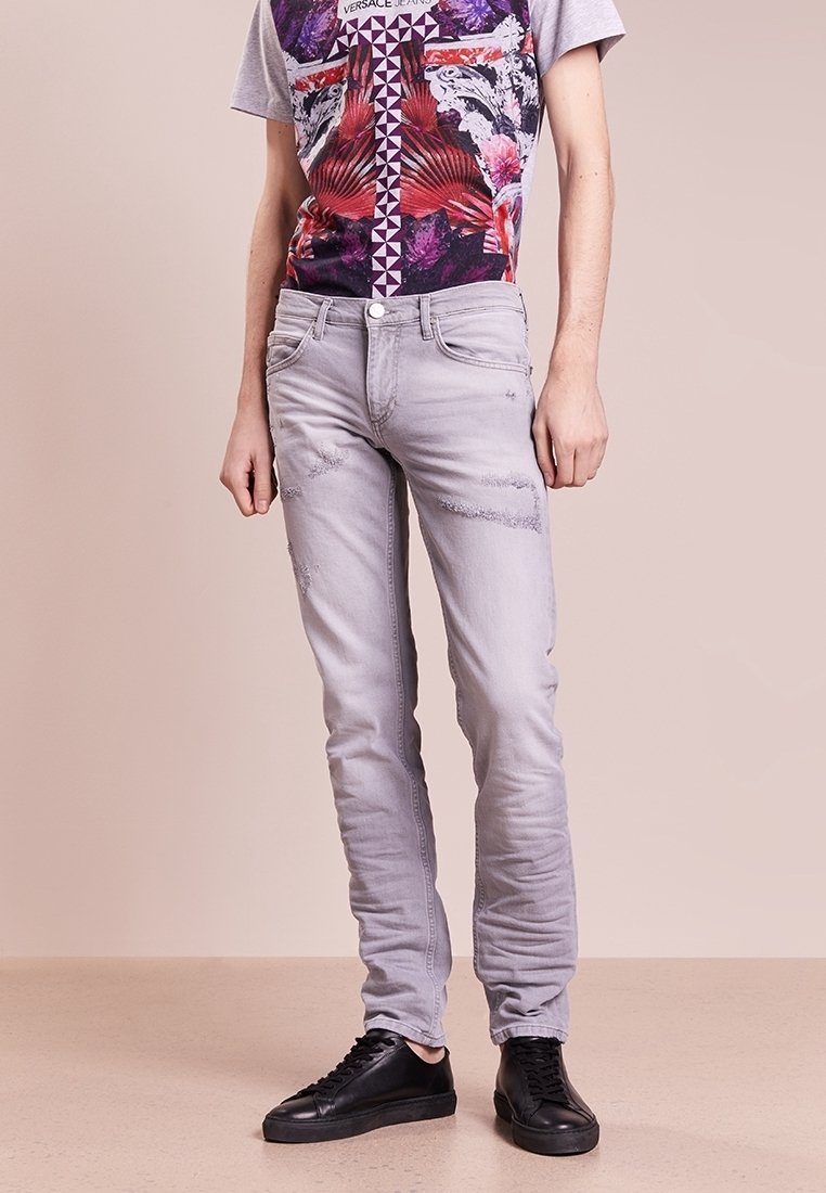 Personne portant un jean gris clair délavé, un t-shirt graphique coloré et des baskets noires, debout sur un fond beige uni.