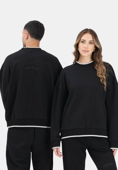 Uomo di spalle e donna di fronte indossano felpe e pantaloni neri coordinati con rifiniture bianche su polsini e orlo.