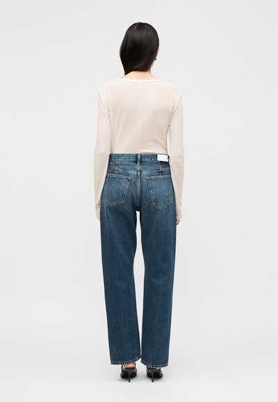 Γυναίκα με μακριά σκουρόχρωμα μαλλιά, που φοράει ένα ανοιχτό μπεζ μακρυμάνικο τοπ, ψηλόμεσα μπλε τζιν και μαύρα τακούνια σανδάλια, στέκεται γυρισμένη προς τα πίσω.