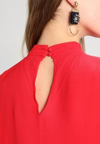 Blusa rossa con dettaglio a chiave sulla schiena e due bottoni, realizzata in tessuto leggero, abbinata a un orecchino decorativo con accenti floreali.