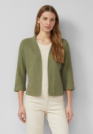 Vrouw met lang lichtbruin haar draagt een olijfgroen gestructureerd open voor jasje met driekwart mouwen over een crèmekleurige top en broek.